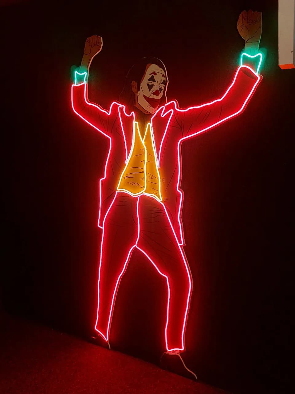 Joker figure panel: red neon outlining the suit, a yellow waistcoat and turquoise cuffs on a dark wall