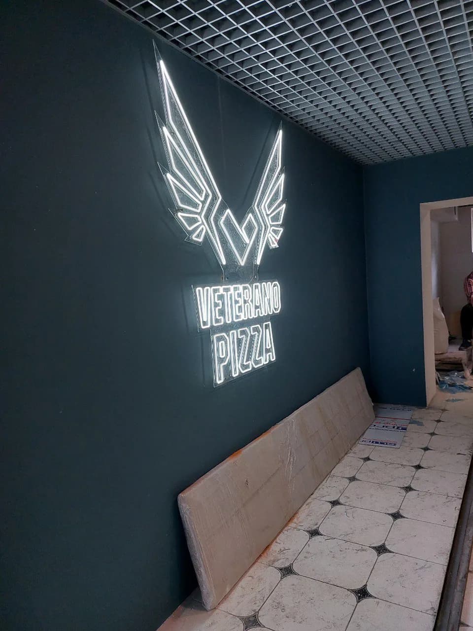 White neon winged emblem and outline VETERANO PIZZA lettering on a dark green dining-room wall
