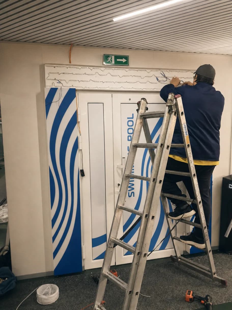 A TheFace installer on a stepladder wiring the illuminated sign above a fitness-club pool entrance